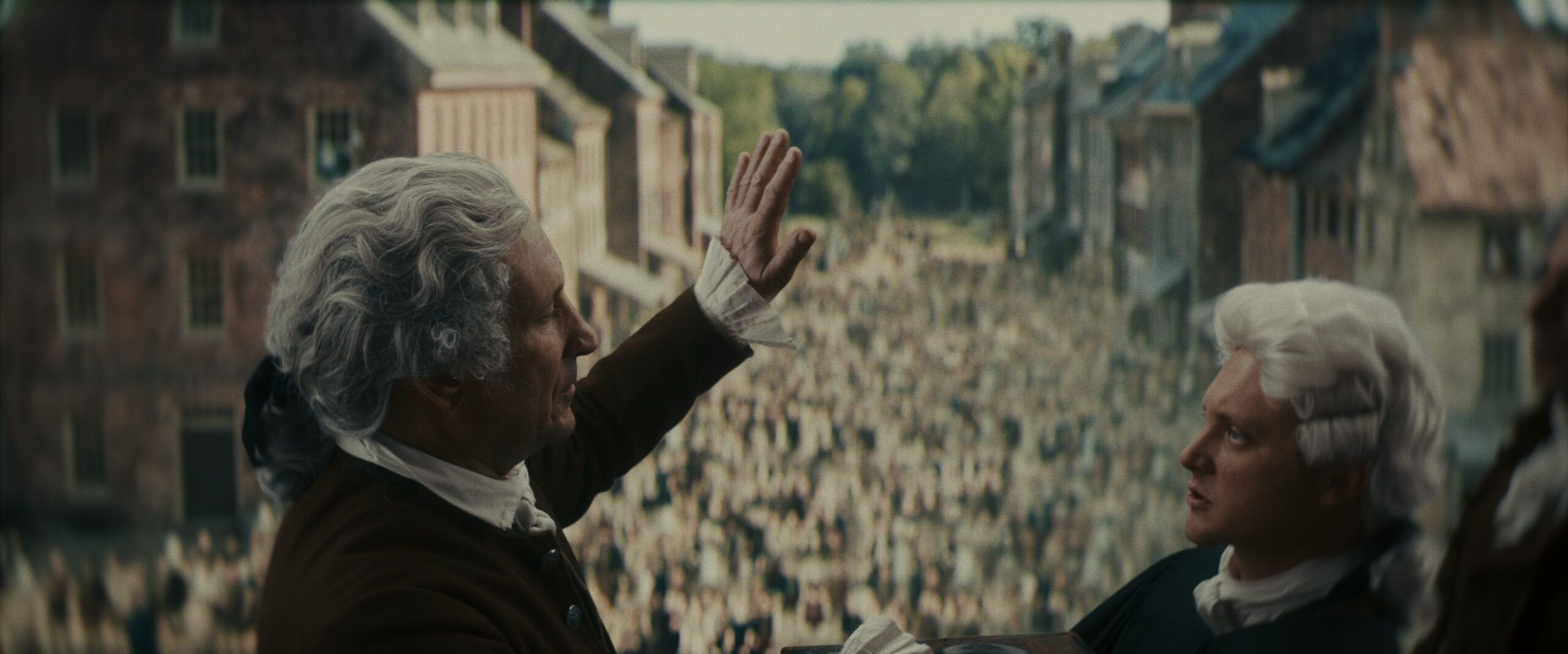 "Two men in 18th-century attire stand on a balcony overlooking a large crowd filling a colonial street lined with brick buildings, with trees visible in the background, suggesting a historical event or public gathering."